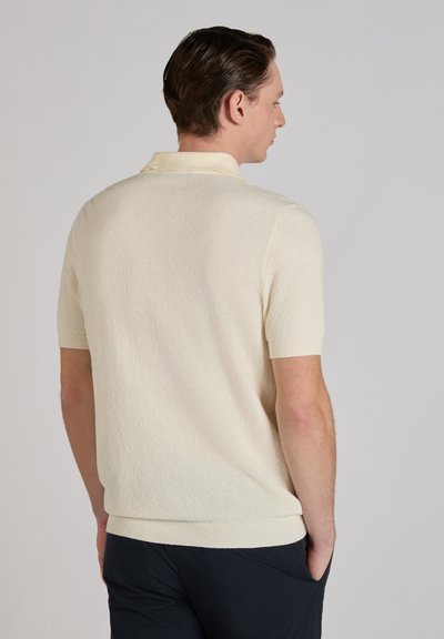 Mężczyzna stoi tyłem, ubrany w kremową, krótkorękawową dzianinową koszulkę polo o fakturowanej strukturze oraz ciemne spodnie, z rękami w kieszeniach, na jednolitym tle.