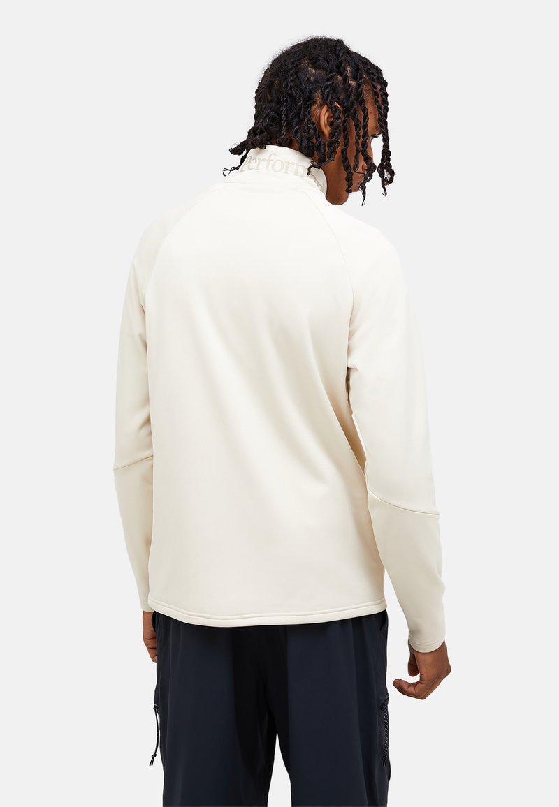 Homme aux cheveux tressés portant un pull crème à manches longues et un pantalon foncé, vu de dos sur un fond uni.