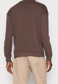 Sudadera marrón con cuello redondo y diseño de hombros caídos, con puños y dobladillo acanalados, fabricada en una tela suave. Pantalones beige claro mostrados a continuación.