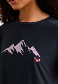 Zwart kortemouwshirt met een roze bergafbeelding op de voorkant en een klein roze logo. Glad textuur met een ronde hals.