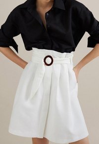 Donna che indossa una camicia nera con bottoni infilata in una gonna bianca a vita alta con pieghe e una cintura in tessuto con una fibbia circolare.