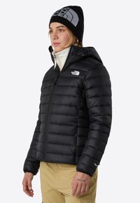 Svart puffjacka med huva, texturerad kviltad design, vit insida och sidofickor med dragkedja. Logotyp på vänster bröst och ärm.