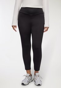 Czarne legginsy mają wysoki pas i dopasowany fason. Noszone z białymi butami sportowymi materiał wygląda na gładki i elastyczny.