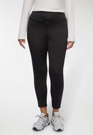 Leggings neri con vita alta e design aderente. Indossati con scarpe da ginnastica bianche, il materiale appare liscio e elasticizzato.