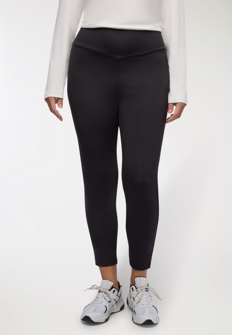 Czarne legginsy mają wysoki pas i dopasowany fason. Noszone z białymi butami sportowymi materiał wygląda na gładki i elastyczny.