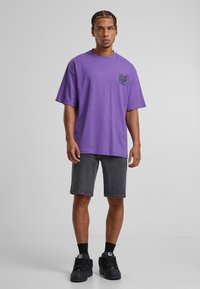 T-shirt en coton violet à manches courtes avec un logo sur la poitrine, associé à un short en denim gris et des chaussures de sport noires.