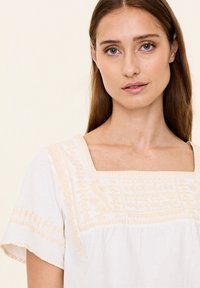 Weiße Bluse mit beiger Stickerei. Hat einen quadratischen Ausschnitt und kurze, ausgestellt Ärmel. Weicher Stoff mit kunstvollen Musterdetails im Oberteil.
