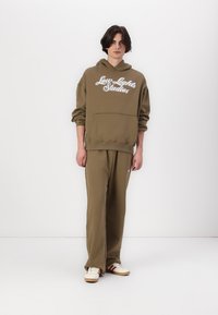 Olivegrüner Kapuzenpullover mit weißem "Low Lights Studios" Text, Kängurutasche, kombiniert mit passenden Hosen und hellen Sneakers.