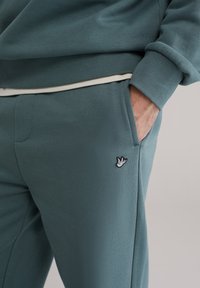 Tirkizne trenirke glatke teksture, s bočnim džepom i malim izvezenim logotipom ptice blizu struka. Rebrasti manžeti i pojas.