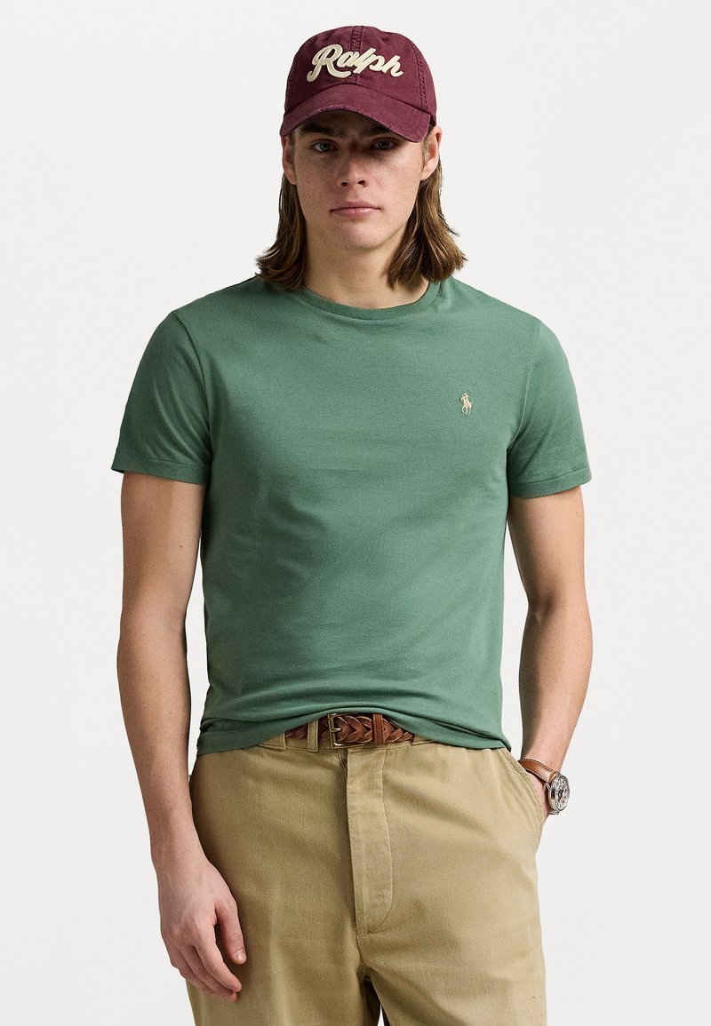 Green cotton t-shirt with a crew neckline, featuring a small embroidered logo. Paired with light beige trousers and a maroon baseball cap.