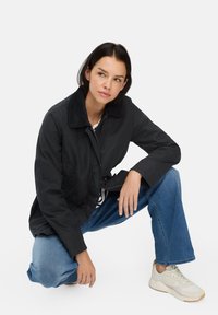 Zwarte jas met een corduroy kraag, voorzakken en een relaxte pasvorm, gecombineerd met lichtblauwe jeans en crèmekleurige sneakers.