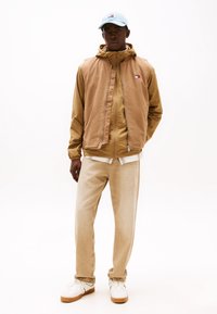 Lichtbruine mouwloze jas over een beige hoodie, gecombineerd met lichtkhaki broeken en witte sneakers. De pet is lichtblauw.