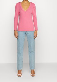 Gestreifter pinker Pullover mit tiefem V-Ausschnitt, langen Ärmeln und Schulterknöpfen, kombiniert mit hellblauen geraden Jeans und weißen Absatzschuhen.