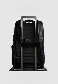 Zwarte rugzak met gevoerde achterkant, met ritssluitingen afsluitbare vakken en verstelbare bandjes, bevestigd aan het handvat van een zilveren tweewielige koffer.