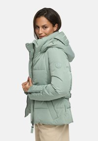 Lichtgroene pufferjack met een hoge kraag en een bevestigde capuchon. Beschikt over ritszakken aan de zijkanten en een ronde logopatch op de mouw.