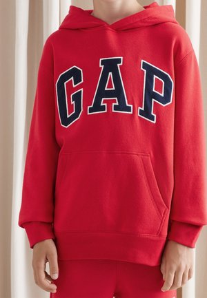 Kind trägt einen roten Hoodie mit großen marineblauen und weißen Buchstaben "GAP" auf der Brust, steht vor einem beigen Vorhang als Hintergrund.