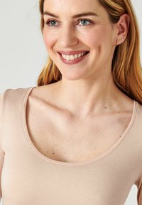 Top rosa chiaro con scollatura a giro e design aderente, texture liscia e dettagli di cucitura sottili. Il modello sfoggia un sorriso naturale e orecchini semplici.