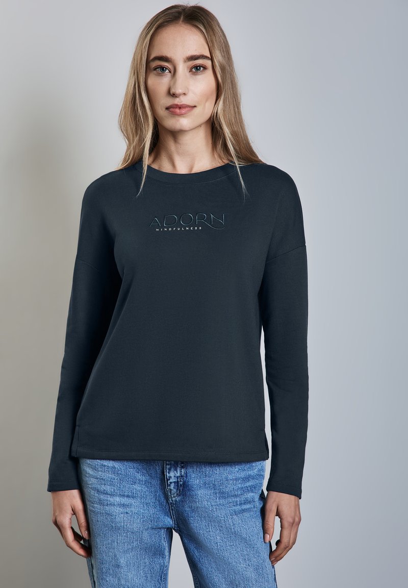 Dunkelgrauer Langarm-Sweatshirt mit der Aufschrift "ADORN MINDFULNESS" auf der Brust, kombiniert mit hellblauen Jeans.