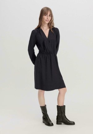 Schwarzes Wickelkleid mit langen Ärmeln, elastischem Taillenbund und knielangem Saum. Getragen mit schwarzen Leder-Ankle-Boots mit Riemen und Reißverschluss.
