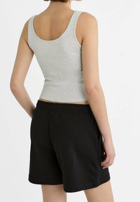 Osoba v šedém bezramínkovém tričku a černých šortkách na elastický pás, zobrazená ze zadní strany na jednoduchém bílém pozadí.