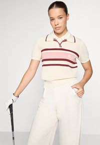 Krémová polo košeľa s marhuľovo-ružovými prúžkami, krátkymi rukávmi a golierom. Kombinovaná s bielymi nohavicami a golfovou rukavicou, drží palicu.