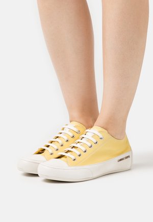 Benen met laag uitgesneden gele leren sneakers met witte veters en zolen, merknaam "Candice Cooper" op de witte zijkant van de zool.