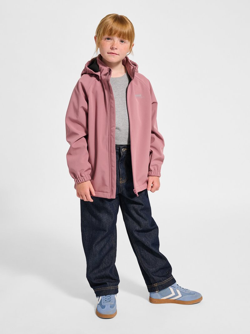 Rødhåret barn står op, iført en pink hættejakke, grå t-shirt, mørke jeans og blå sneakers med hvide striber.