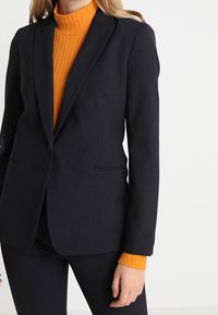 Svart skräddarsydd kavaj med två framfickor, notched lapels och en slät textur, överlagd på en orange ribbad polotröja.