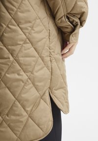 Beige vadderad kappa med diamantstickning på en persons arm och hand, visar sidoflik och tryckknapp på manschetten.