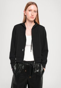 Svart zip-up hettegenser med en tettsittende design, med snor i hetta og sølvfarget metall. Bærer den over en hvit topp og svarte skinnbukser.