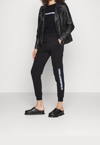 Chaqueta de motorista de cuero negro sobre una camiseta negra, combinada con pantalones jogger negros con letras azules y zapatillas negras sin cordones.