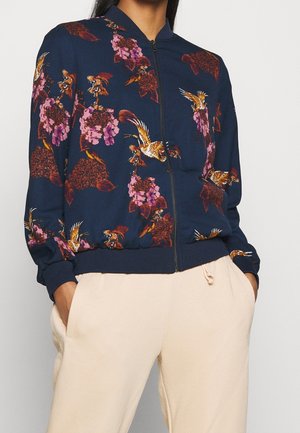 Marineblauw jack met ritssluiting en bloemen- en vogelprint, gedragen met crèmekleurige broek met trekkoord, handen in de zakken gestoken.