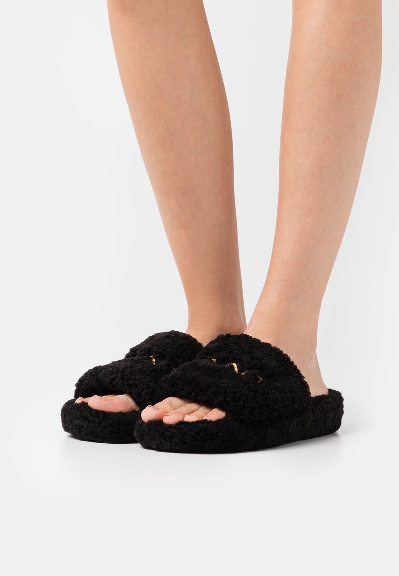 Pieds portant des chaussons noirs duveteux à bout ouvert avec une semelle épaisse, présentés sur un fond blanc uni.