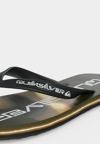 Κοντινό ενός μαύρου σαγιονάρου Quiksilver με λευκό λογότυπο στο λουράκι και τη σόλα, υφή στη σόλα και λεπτομέρεια στρωμάτων στη μεσαία σόλα.