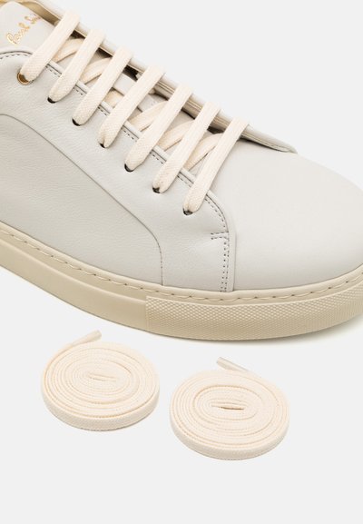 Zapatilla blanca de cuero con suela de goma beige, con cordones planos beige y una textura suave. Se incluyen dos cordones adicionales a continuación.