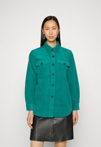 Tee-grüne Cordhemd mit durchgehender Knopfleiste, zwei Brusttaschen und langen Ärmeln. Getragen über einem schwarzen Lederrock.