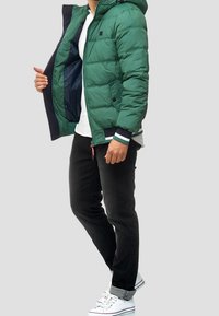 Giacca piumino verde con cappuccio, chiusura a zip, due tasche laterali, polsini a coste e un interno blu a contrasto. Abbinata a jeans neri e sneakers.