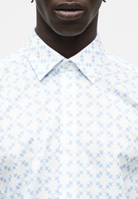 Gros plan d'une personne portant une chemise blanche à boutons avec un motif géométrique floral bleu clair et un col pointu.