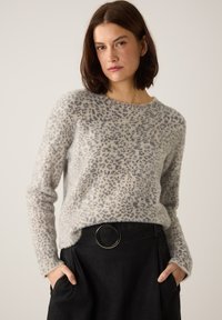Hellgrauer, flauschiger Pullover mit einem Leopardenmuster in dunkleren Grautönen, rundem Ausschnitt und langen Ärmeln. Kombiniert mit einem schwarzen Rock, der einen großen silbernen Ringgürtel hat.