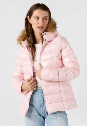 Donna che indossa una giacca a vento rosa chiaro con cappuccio foderato di pelliccia, una camicia bianca e jeans azzurri, mentre sistema la cerniera della giacca.