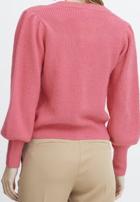 Suéter de punto rosa con textura acanalada, mangas abullonadas y cuello redondo, combinado con pantalones beige que muestran un corte relajado.