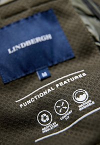 Mörkgrön tygetik med "LINDENBERGH"-etikett i vit text, ikoner för återvunnen isolering, bekväm passform och tvättbarhet i maskin.