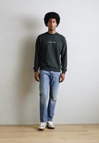 Sweatshirt vert foncé avec du texte blanc, jean bleu et baskets rayées vertes. Le mannequin se tient sur un sol en bois devant un fond uni.