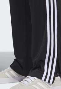 Svarta träningsbyxor med vita ränder på sidorna, med en lös passform och dragkedjor vid anklarna. Matchas med ljusgrå sneakers.