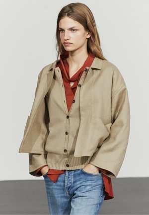 Young woman with long hair wears beige jacket over tan buttoned cardigan and rust red shirt, paired with blue jeans, hands in pockets.