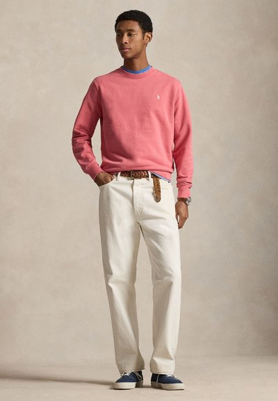 Άνδρας που φοράει κοραλλί ροζ φούτερ, λευκό παντελόνι, καφέ πλεκτή ζώνη, μπλε αθλητικά παπούτσια και ρολόι, στέκεται με το ένα χέρι στην τσέπη.