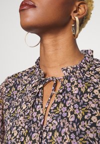 Blusa floral con fondo oscuro, cuello con volantes y detalle de lazo. Presenta patrones florales en púrpura y amarillo. Pendientes de aro dorados.