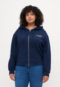 Sudadera con cremallera en color azul marino, con bolsillos frontales, capucha con cordón y logo bordado. Puños y dobladillo acanalados. Combinada con jeans azules.