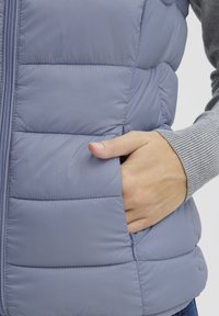 Helllila Puffer-Weste mit horizontalen Steppnähten, einem durchgehenden Reißverschluss und Seitentasche. Unter der Weste befindet sich ein grauer, gerippter Pullover.