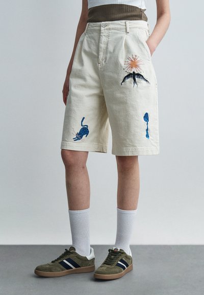Shorts in cotone color crema, decorati con un ricamo blu raffigurante animali e sole. Abbinati a scarpe da ginnastica grigie e calzini bianchi. Vestibilità casual.